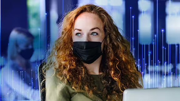 A woman with a mask sitting on a chair with laptop and looking aside
