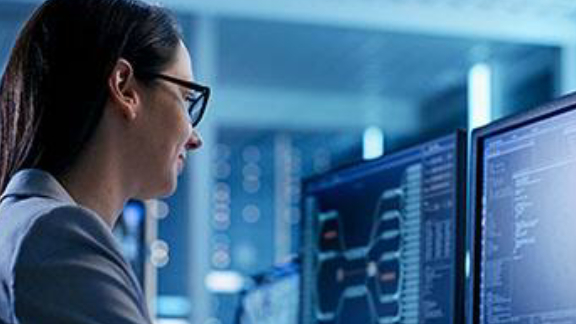 A woman wearing glasses, looking at two monitors displaying code