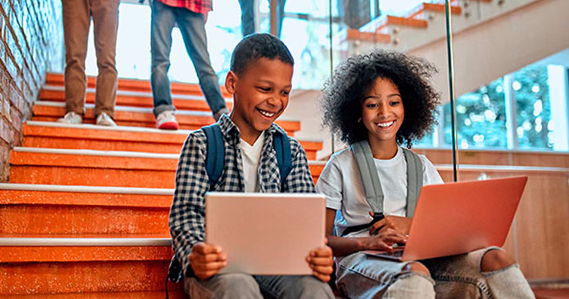 Two children sitting on the stairs and smiling while working on a laptop