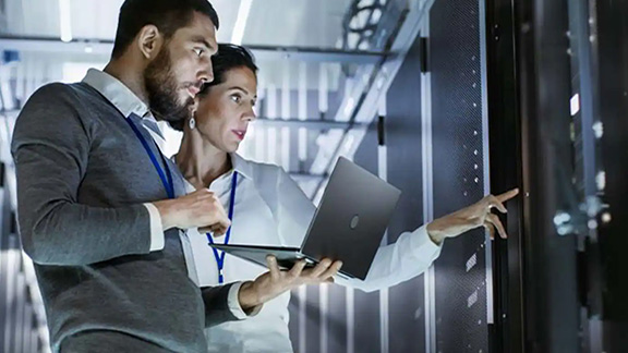 A man holding a laptop. standing next to a woman pointing at a server unit