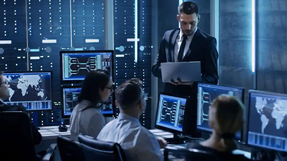 A group of people seated around desktops, with a man standing and holding a laptop, in an office