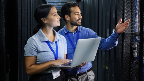 A male technician and a woman holding a laptop talking while walking next to server racks in a data center