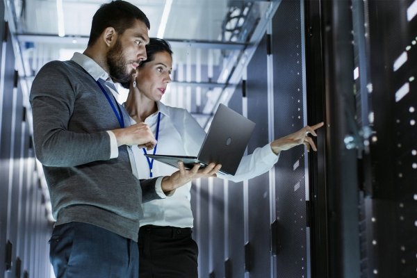 A man holding a laptop standing next to a woman pointing at a control panel