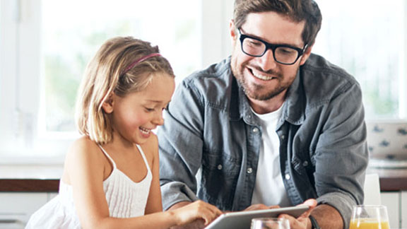 A man and his young daughter at the table, looking at a tablet and smiling
