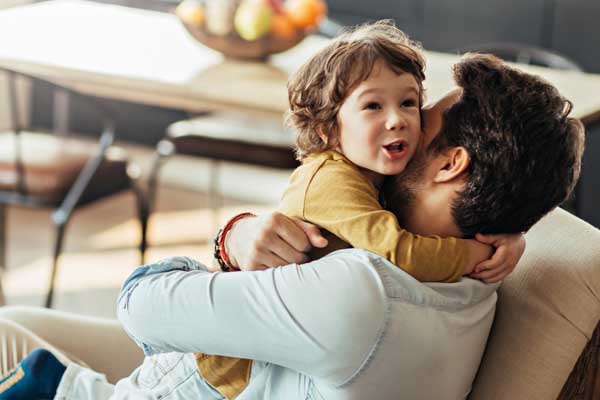 A child smiling and hugging his father around the neck on a living room couch