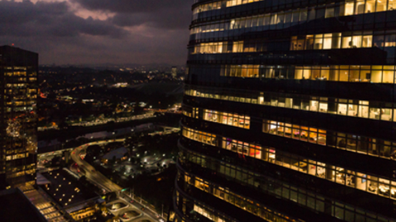 A corporate building lit up at dusk, with a cityscape behind