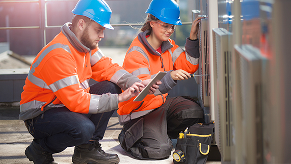 Two maintenance workers kneeling before a device, while one scrolls on a tablet