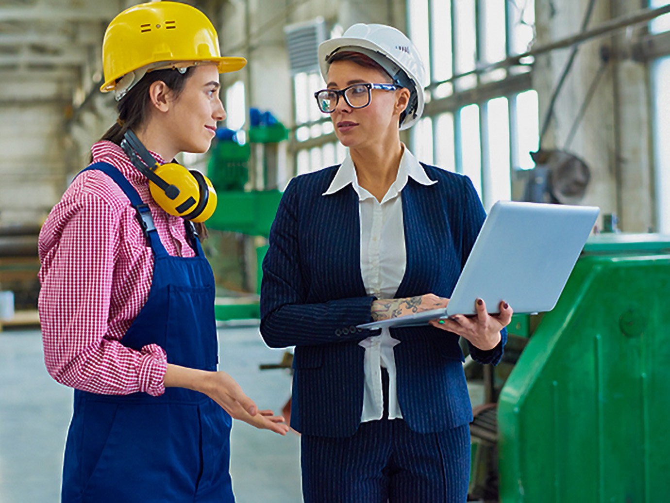 Two women conversing in a factory with one woman holding a laptop